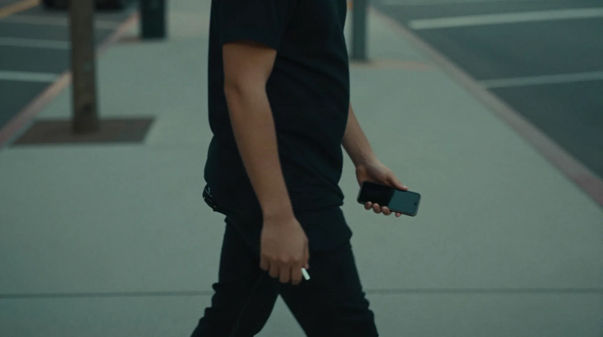 Person walking along a city sidewalk at dusk with a phone in hand and wireless earbuds in