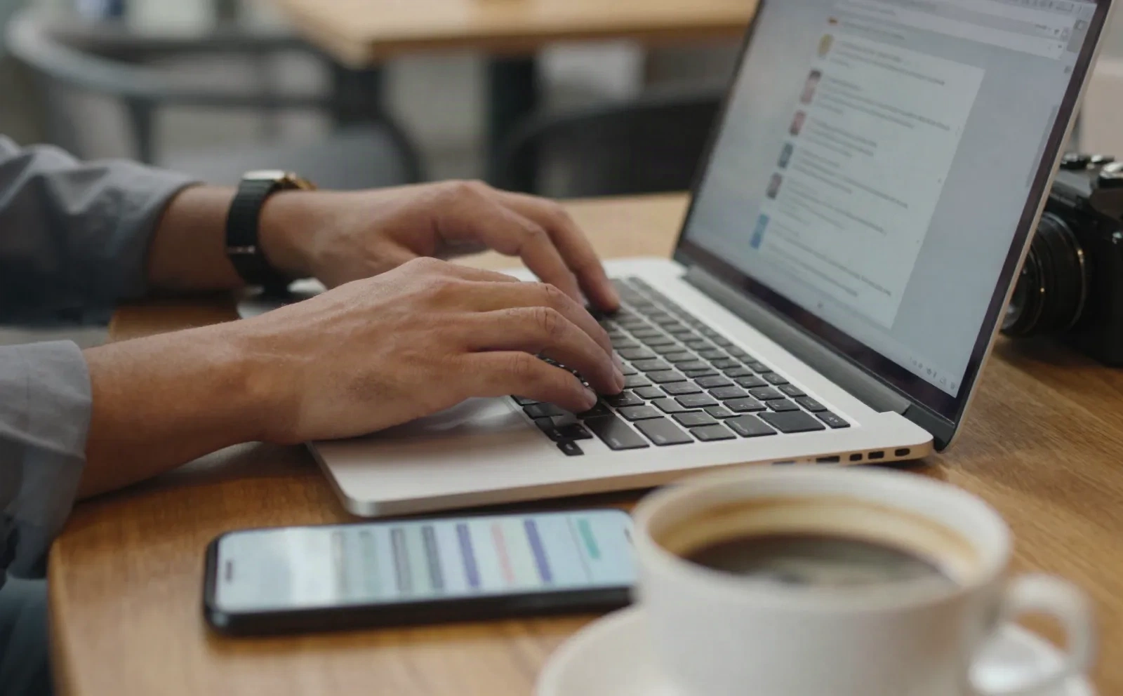Hände tippen auf Laptop in Café, Telefon zeigt Nachrichten-Benachrichtigungen, Kaffee daneben